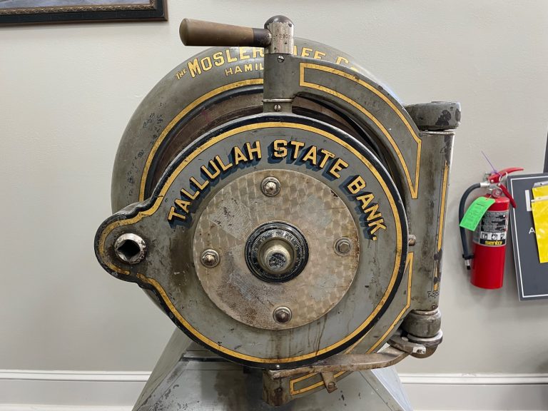 Round grey screw door of a Mosler cannonball safe lettered Tallulah State Bank in gold, with a central combination dial, a crank handle on top and the door hung on a swinging crane